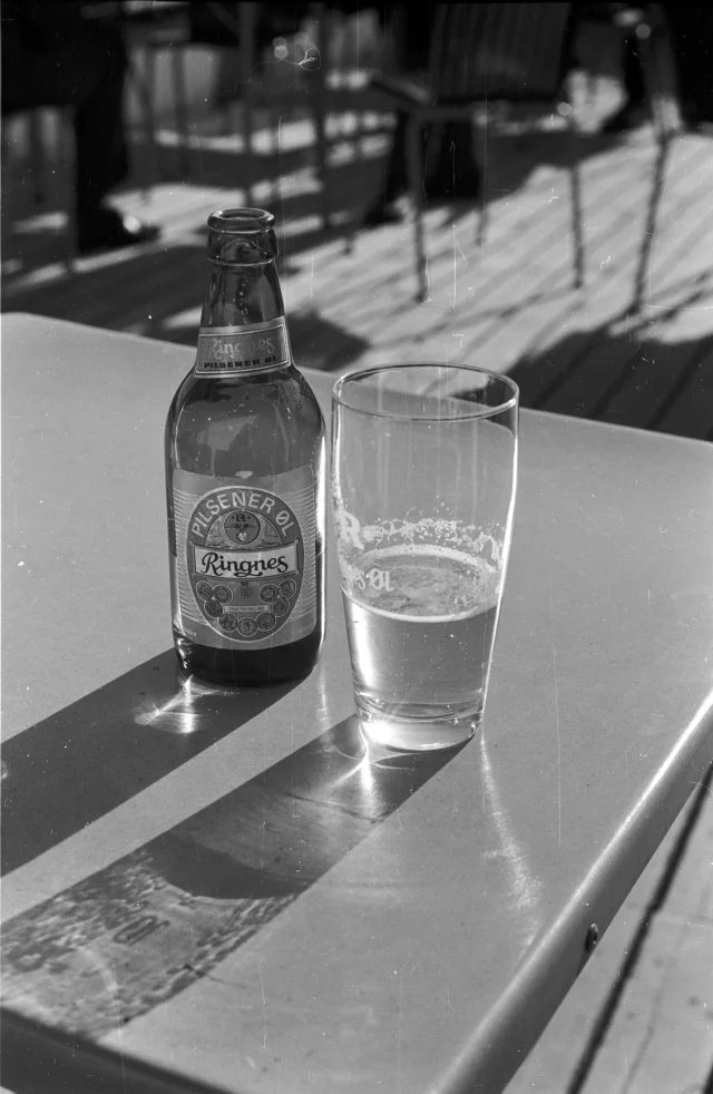 Sort-hvitt-bilde av en ølflaske med Ringnes Pils og et halvfullt ølglass, i solskinn på et bord med uteservering.